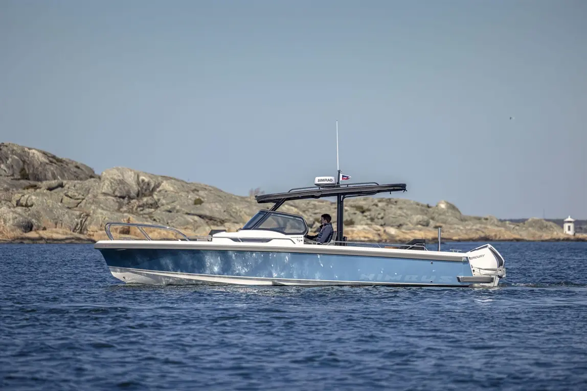 A Nimbus boat navigating calm waters, with rocky shoreline in the background and a driver at the helm, visible SIMRAD branding.